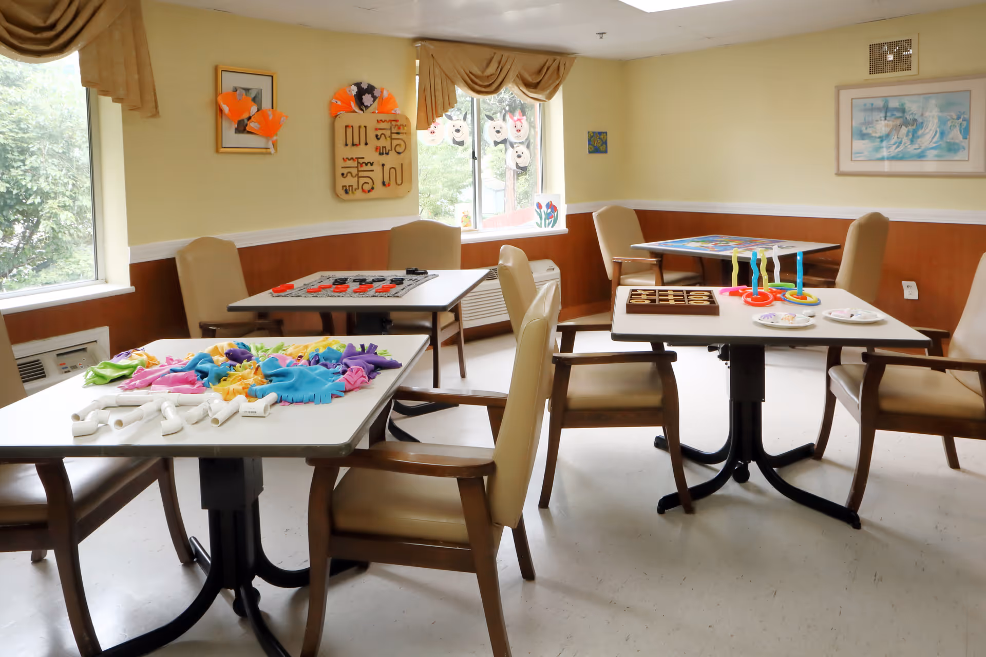 Interior activity room with tables, chairs, crafts, and windows in a senior facility.