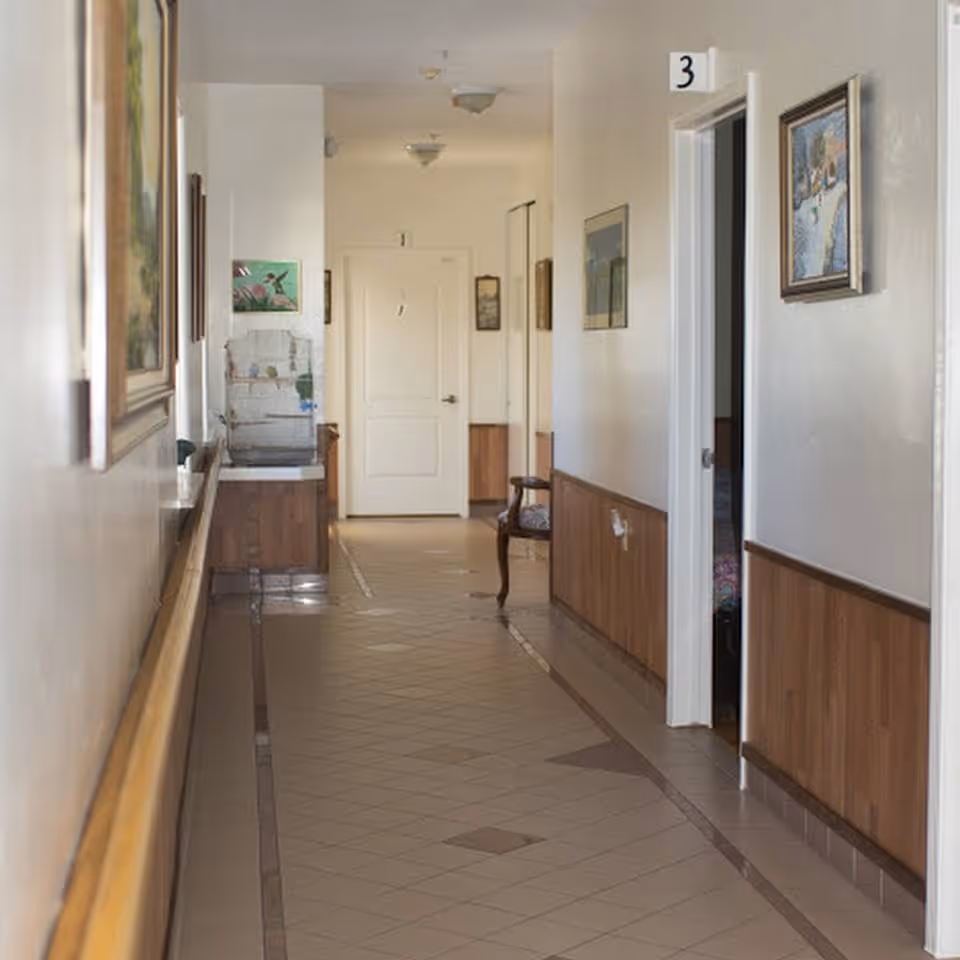 Long tiled hallway inside an assisted living facility with numbered doorways, framed paintings, a chair and handrail.