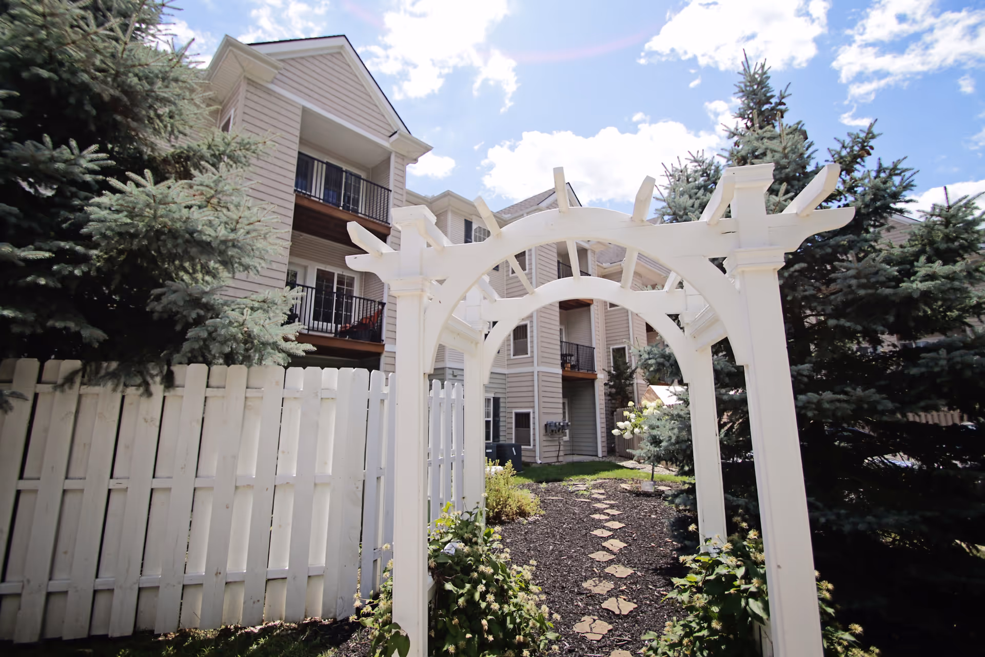 White wooden archway frames a stone path toward a beige apartment building, surrounded by evergreens.