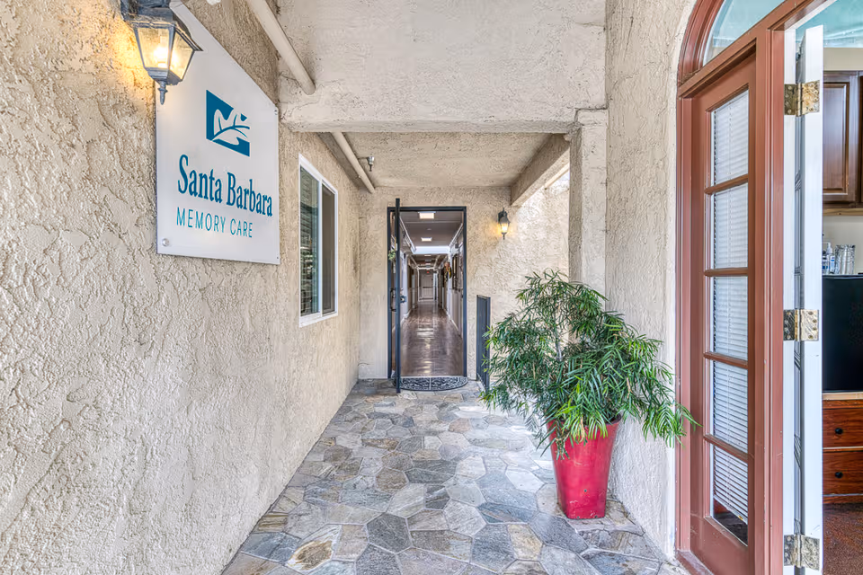 Exterior covered entrance to Santa Barbara Memory Care with sign and potted plant.