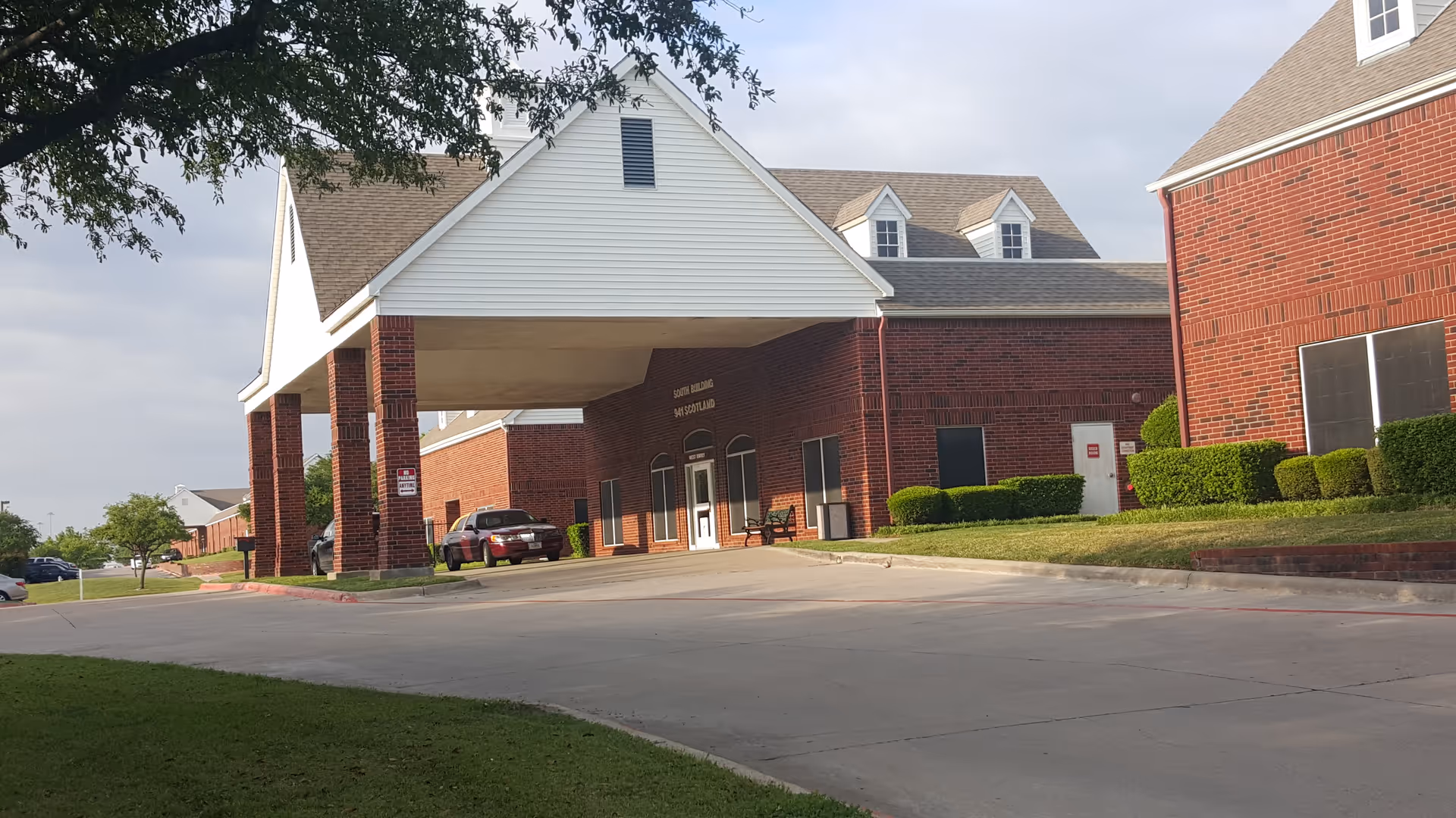 Brick senior living facility with a covered entrance, driveway, and manicured hedges.