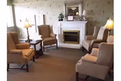 A cozy senior living lounge featuring beige armchairs, lamps, and a fireplace around a mantle.