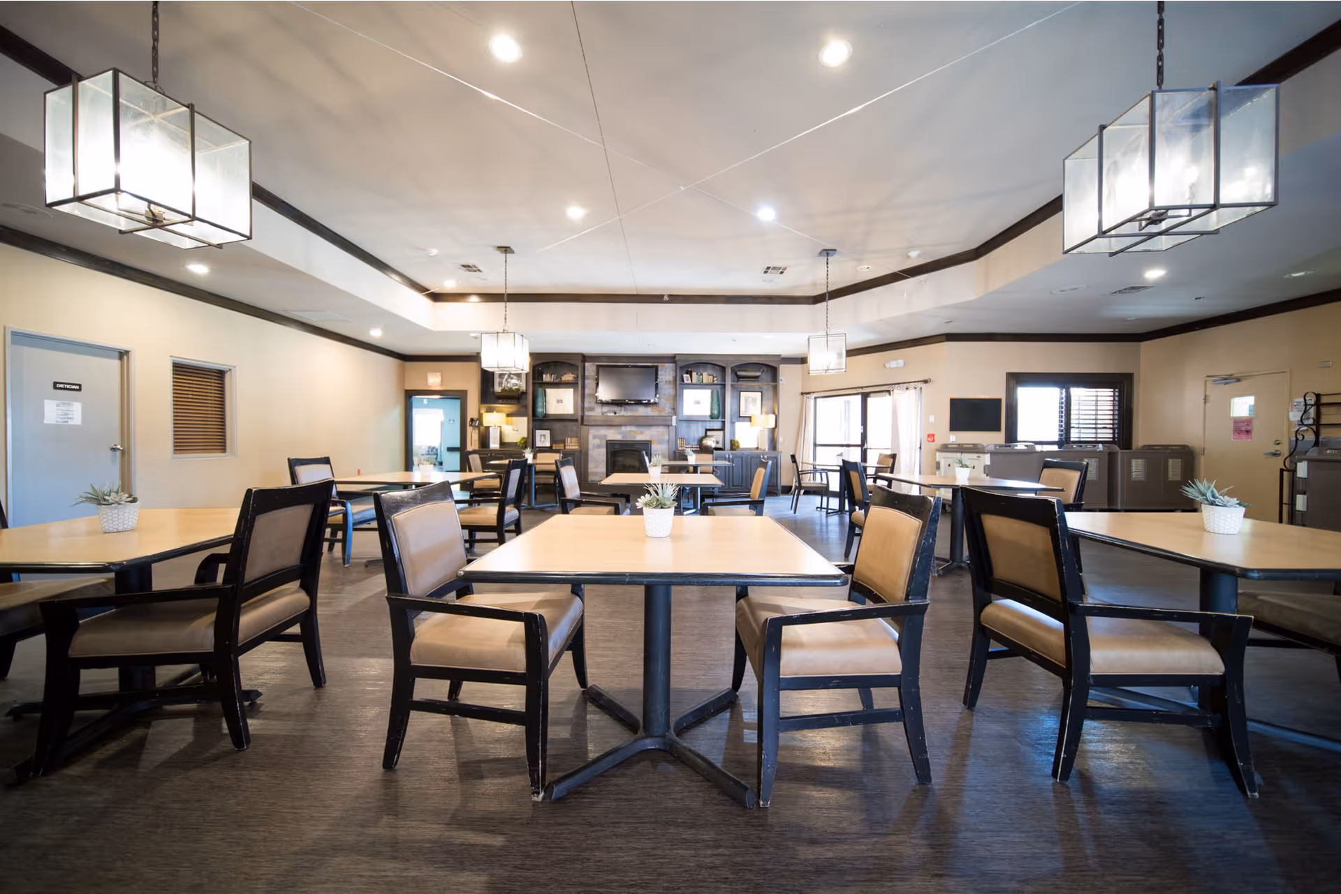 A spacious dining room with multiple square tables and cushioned chairs arranged neatly. The room features modern hanging light fixtures, a fireplace with a TV mounted above it, and large windows allowing natural light to enter. Small potted plants are placed on the tables.