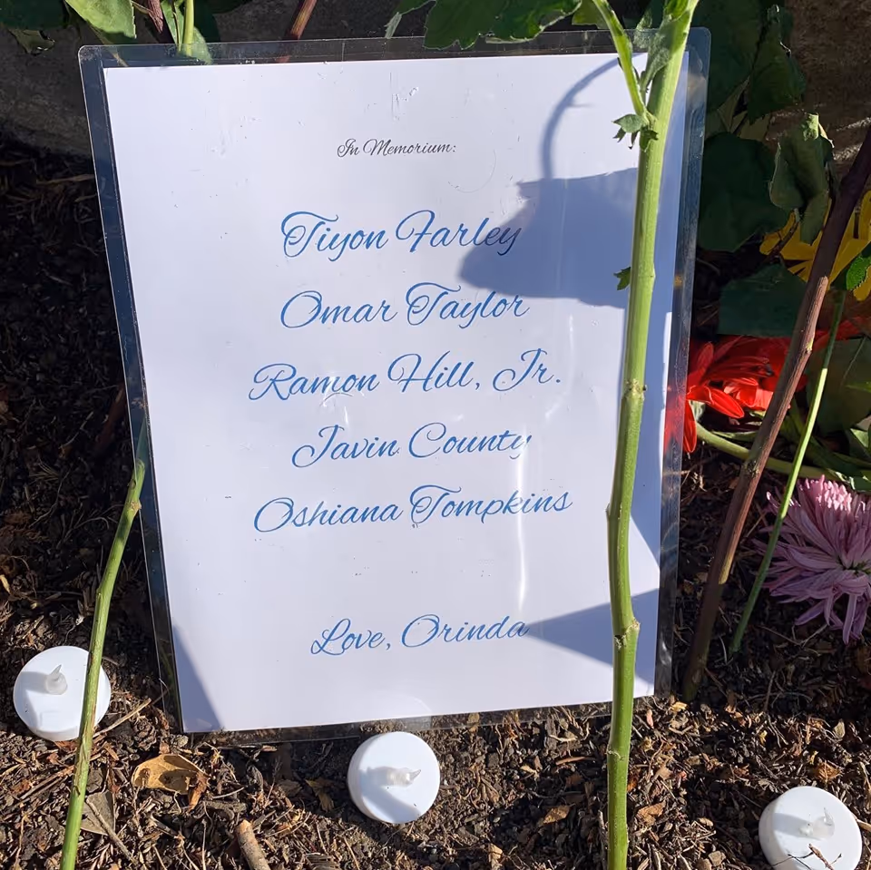 Outdoor memorial plaque with names inscribed in cursive, surrounded by plants and candles.