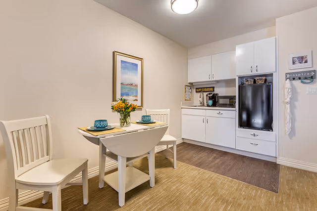 Bright dining nook with white table, two chairs, flowers, and a compact kitchen in the background.