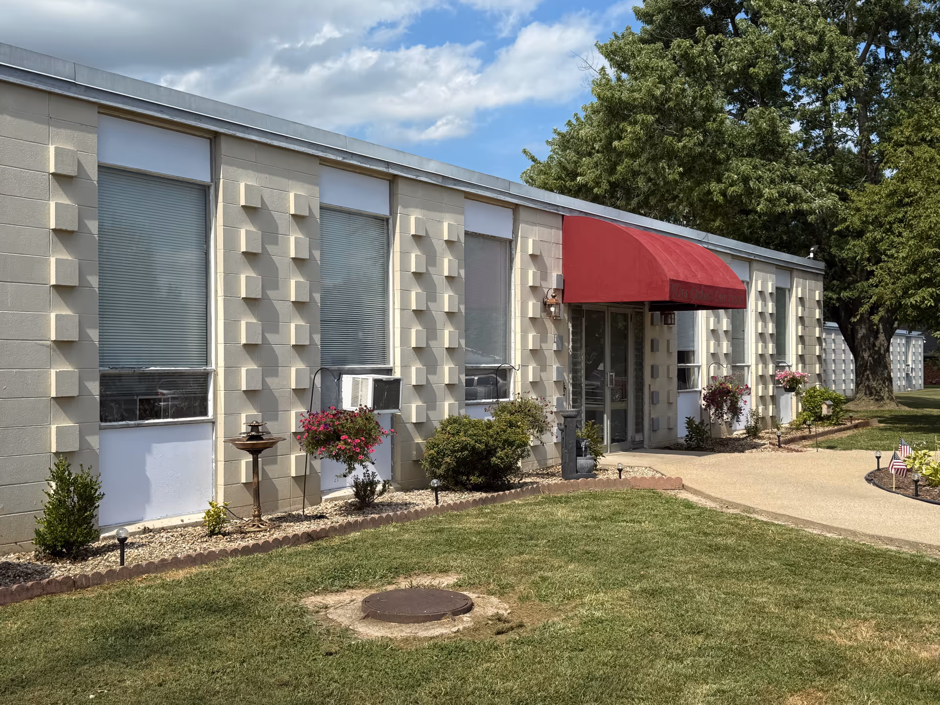 One-story beige masonry building with large windows, a red awning over the glass entrance, and a landscaped lawn with shrubs and trees.