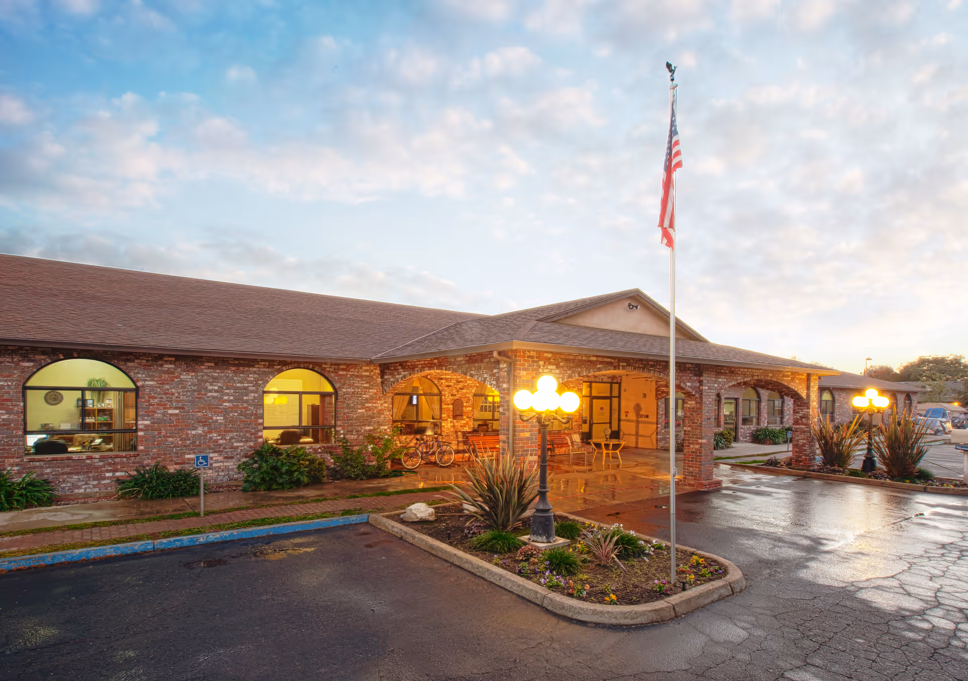 Exterior view of Westgate Gardens Care Center at dusk, showing a single-story brick building with arched windows and a covered entrance. There are lit street lamps, an American flag on a flagpole, some landscaping with plants and flowers, and a wet driveway with parking spaces.