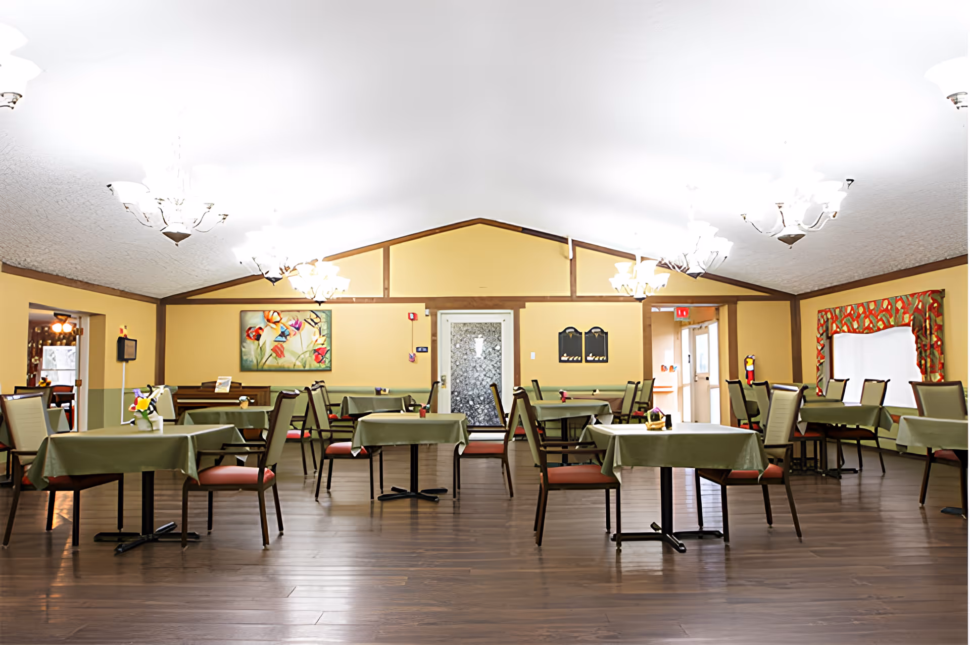 Spacious dining room with multiple tables covered in green tablecloths and chairs beneath chandeliers.