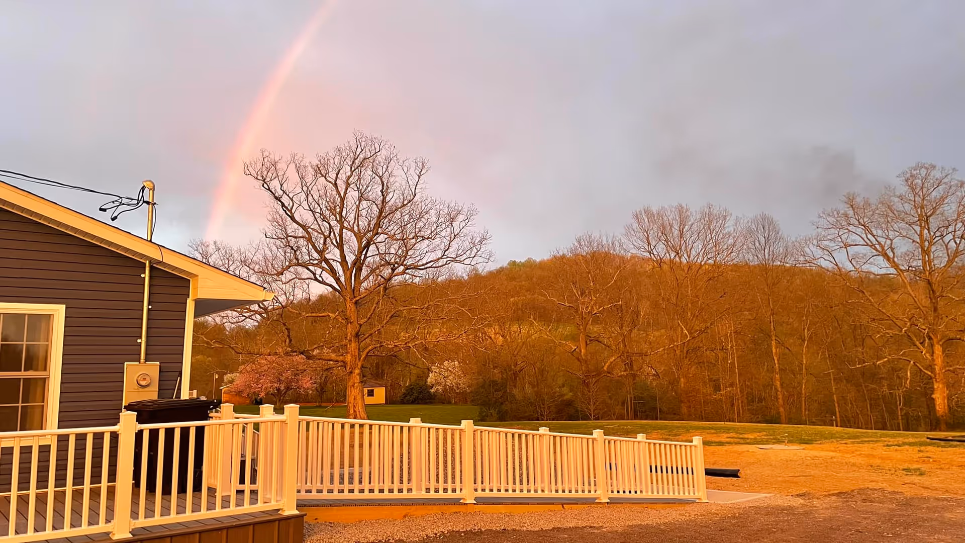 Blue building with white railing deck, rainbow over leafless trees.