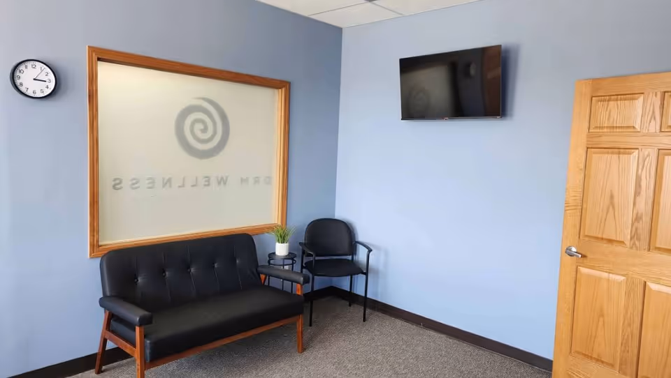 Blue-walled waiting area with sofa, chairs, small plant, clock, and mounted TV.