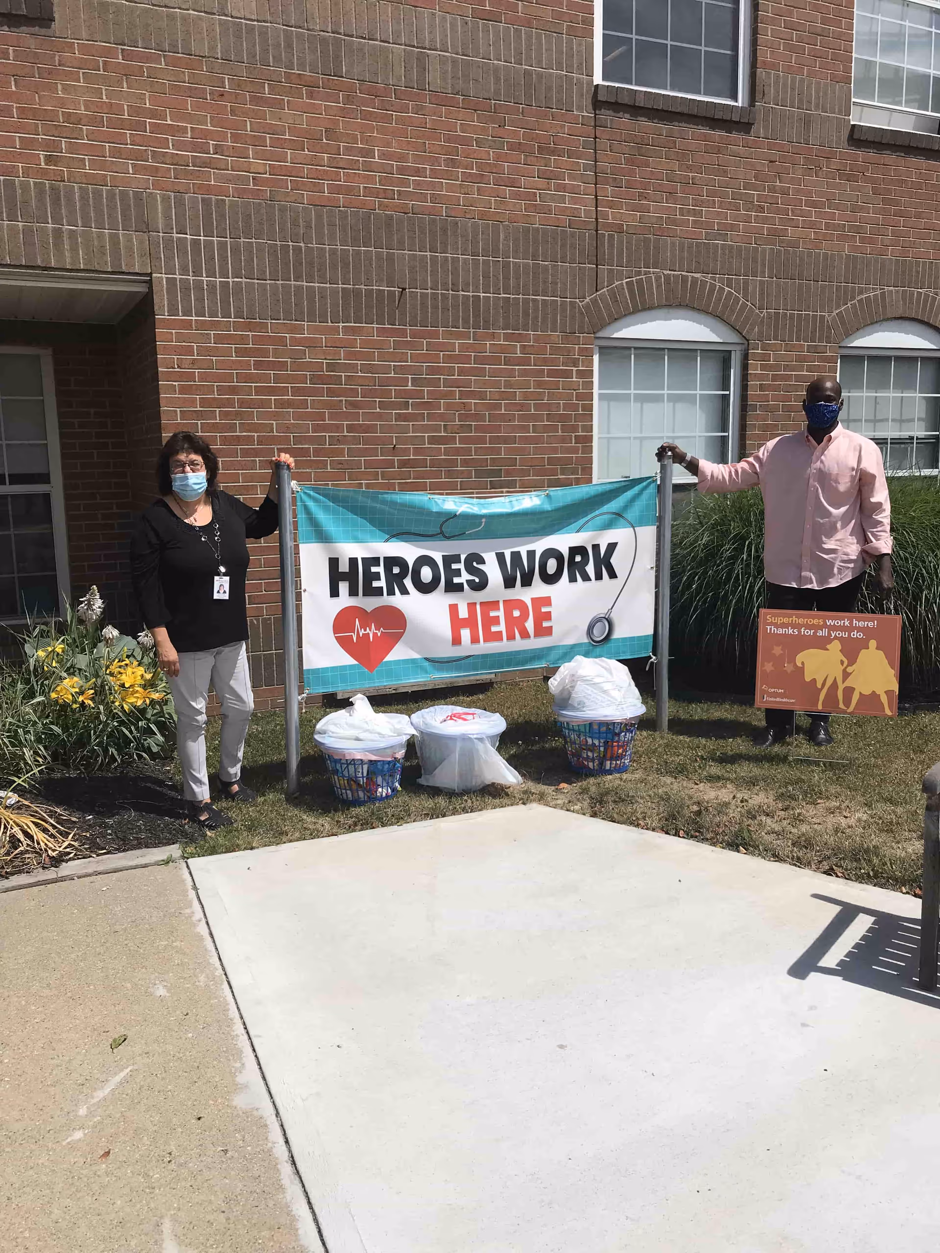 Two people wear masks beside a banner reading Heroes Work Here outside a brick facility.