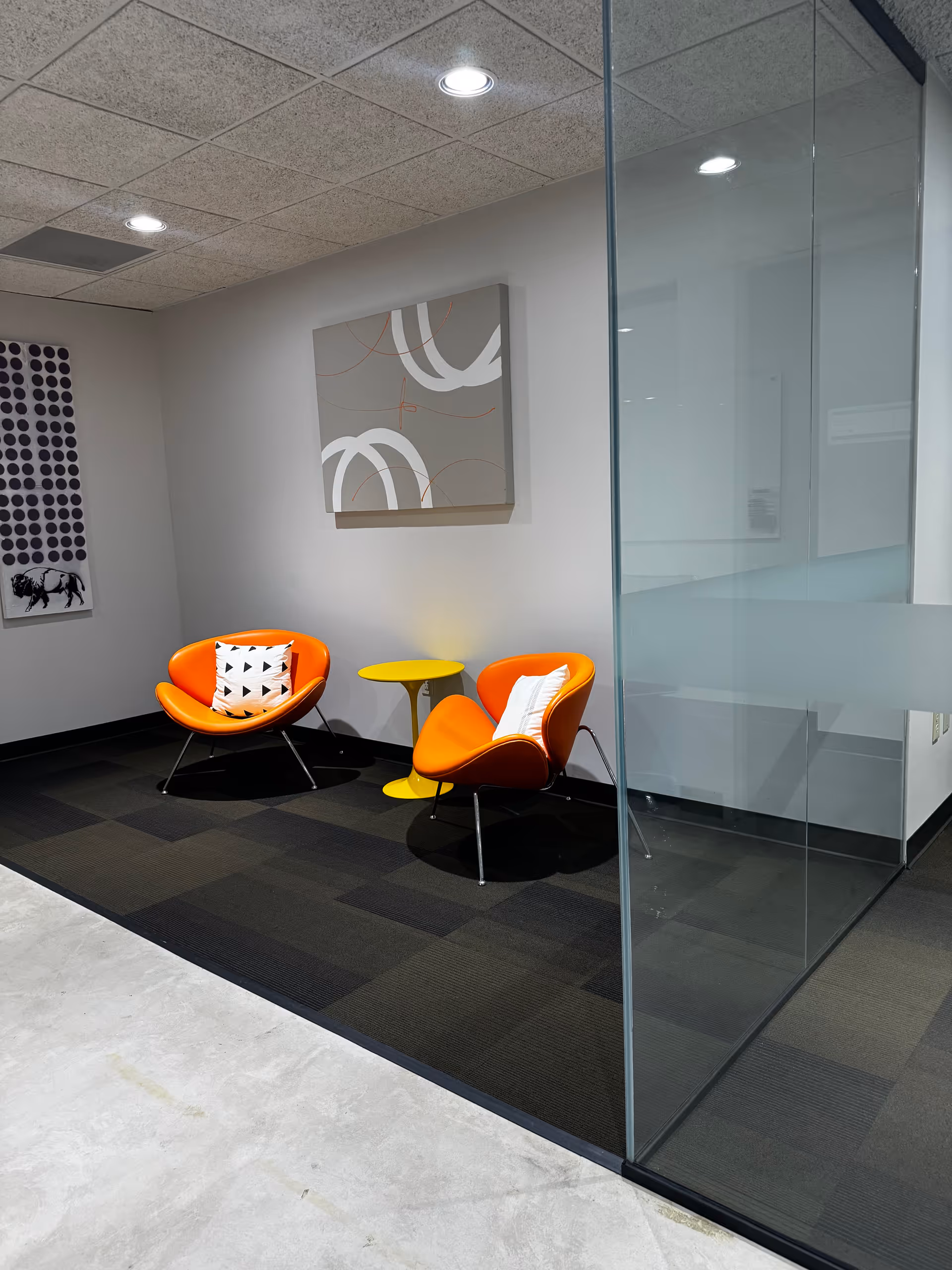 Two orange lounge chairs with a yellow side table in a glass-walled lobby.