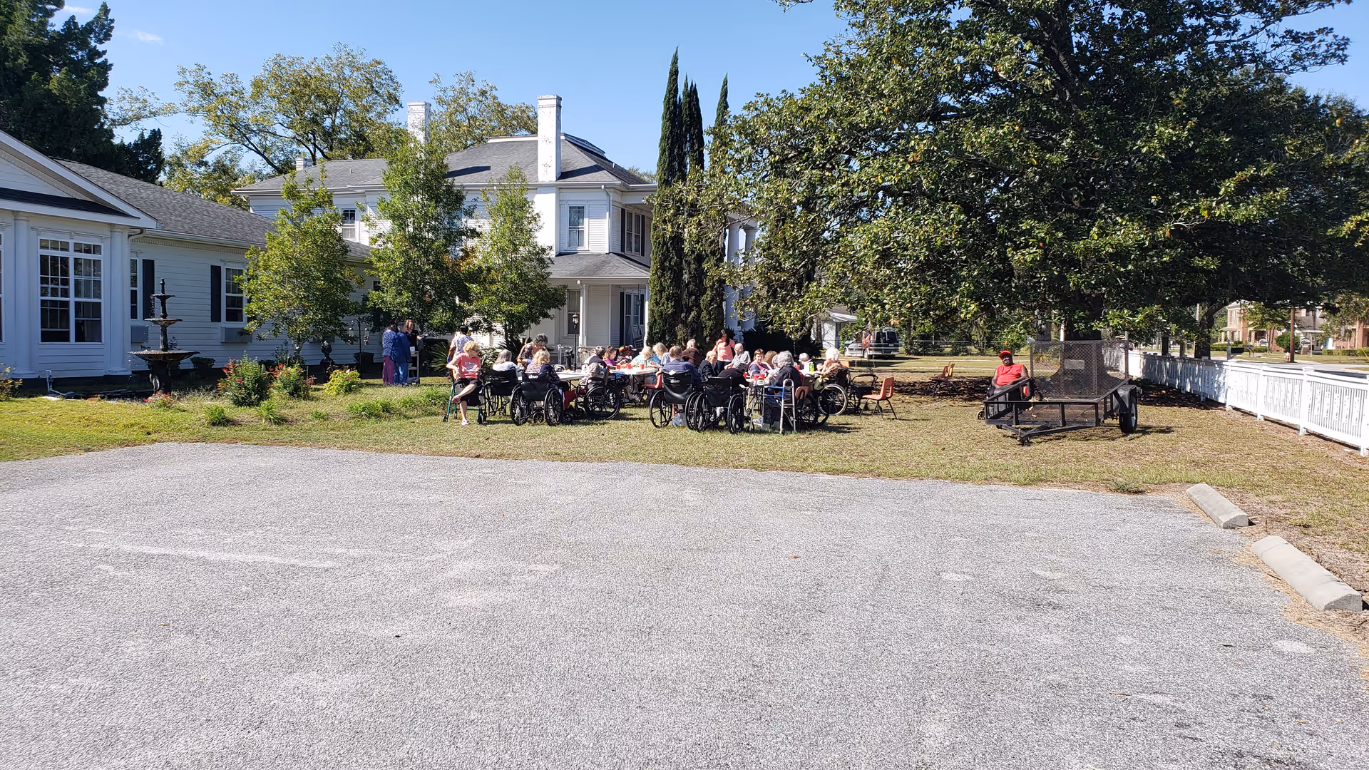 Seniors in wheelchairs and chairs gather outside a white building with trees.
