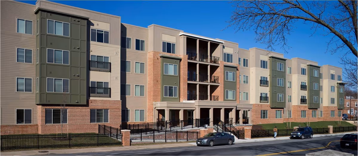 Front exterior of a multi-story senior living building with brick and siding facade, balconies, and cars parked on the street.