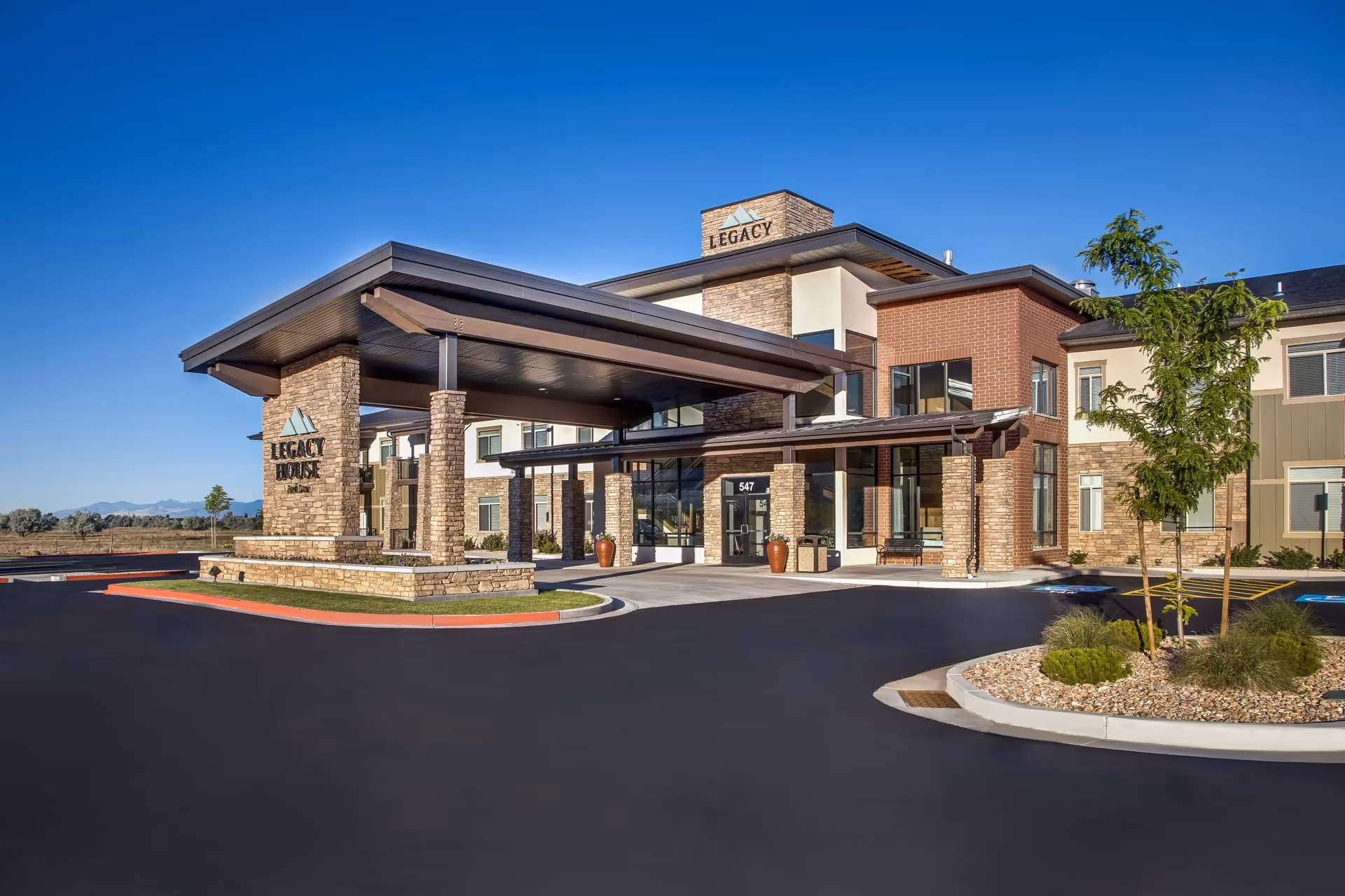 Exterior view of Legacy House of Park Lane, a modern senior living facility with stone and brick facade, large covered entrance, and clear blue sky in the background.