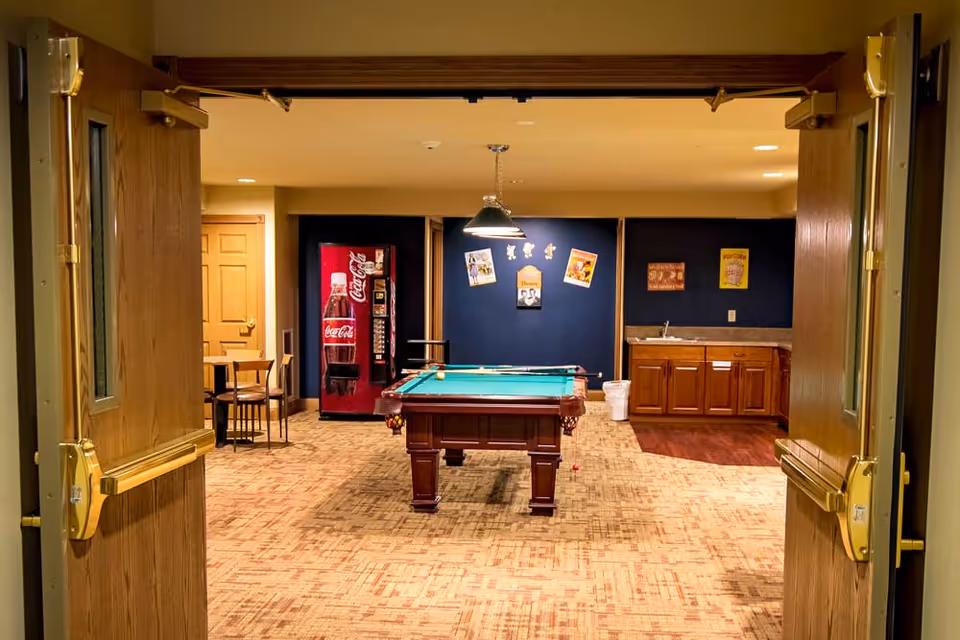 View through open double doors into a recreational room featuring a pool table in the center, a Coca-Cola vending machine against the back wall, a small seating area with tables and chairs to the left, and a kitchenette with wooden cabinets and a sink to the right. The walls are decorated with various posters and the room is warmly lit.
