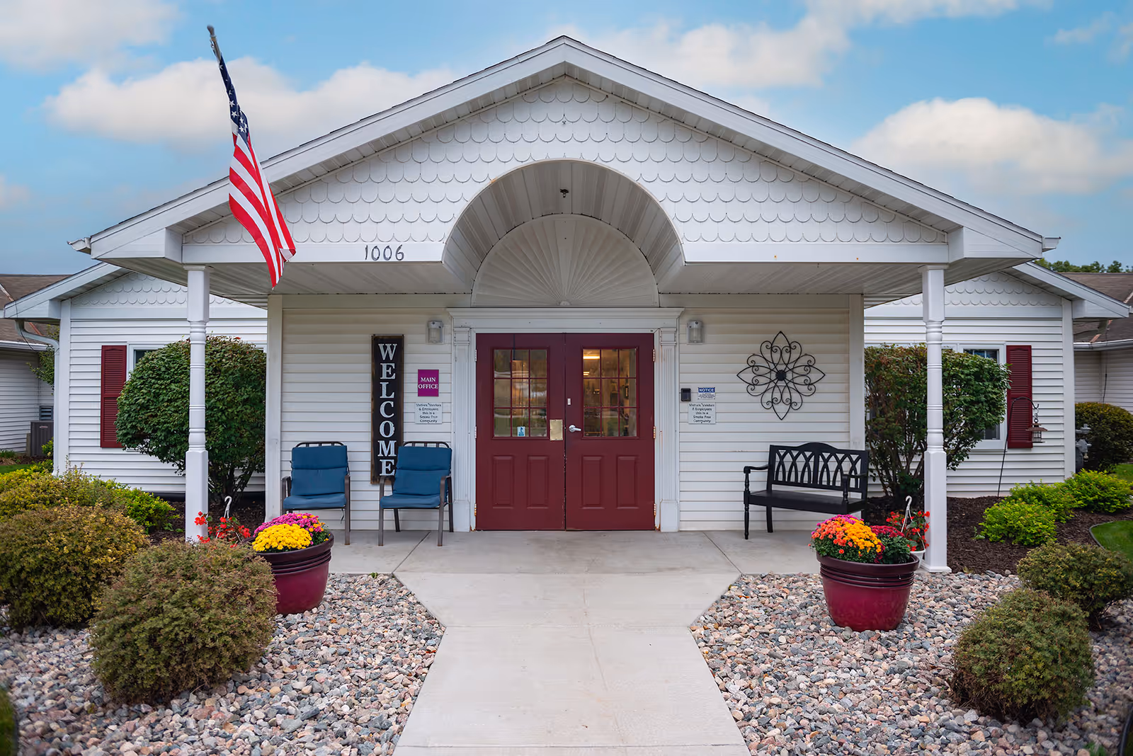Front of a white senior living facility with a red door, flag, and benches.