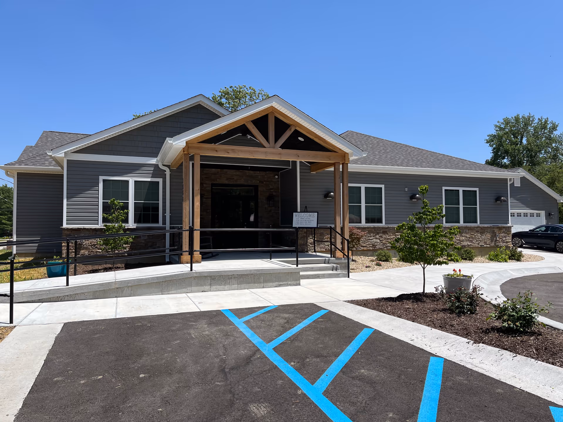 Front exterior of a gray senior living facility with a wooden entrance canopy and handicap markings