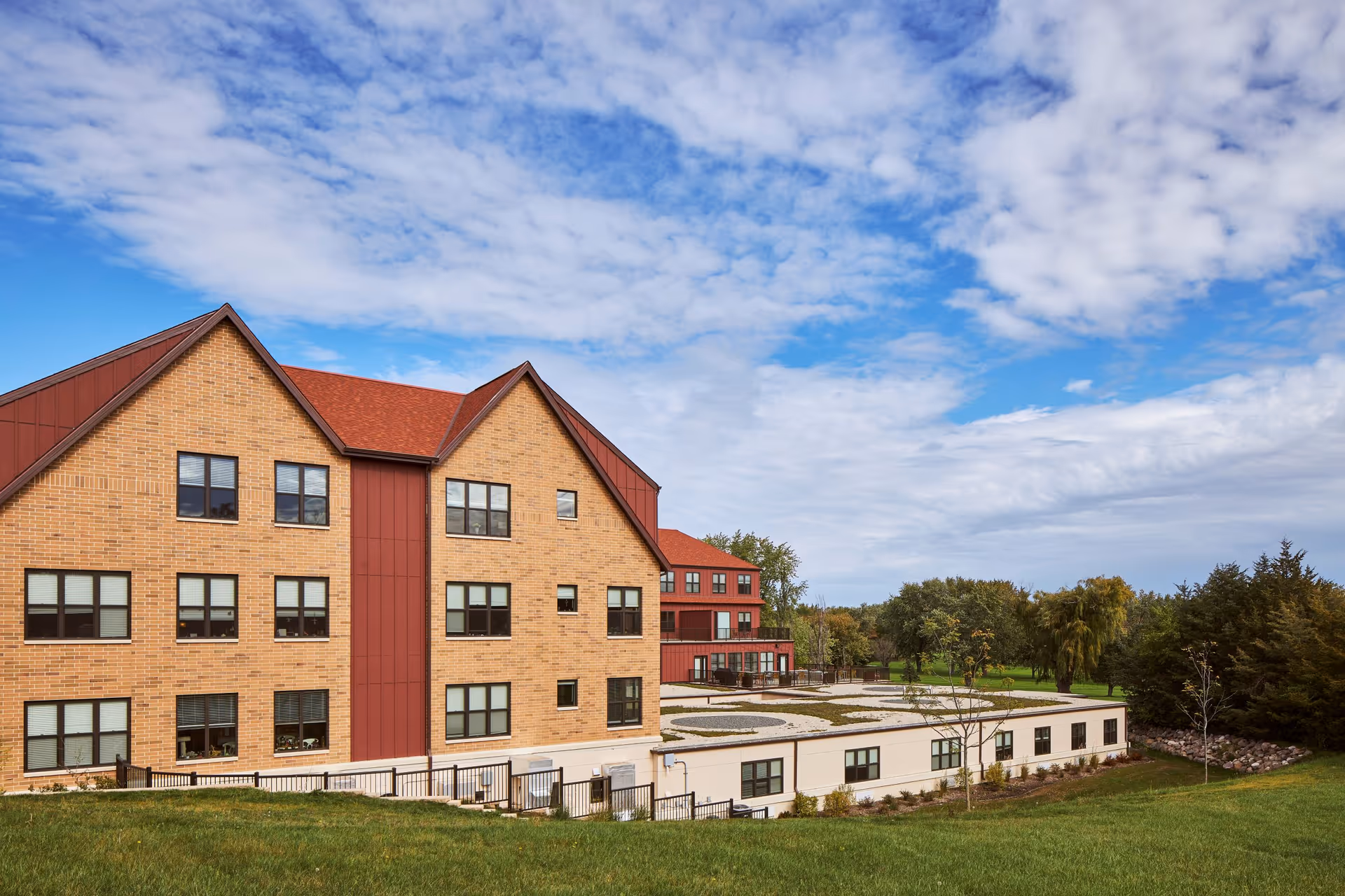 Exterior view of a multi-story senior living facility building with brick and red paneling under a partly cloudy blue sky, surrounded by green grass and trees.
