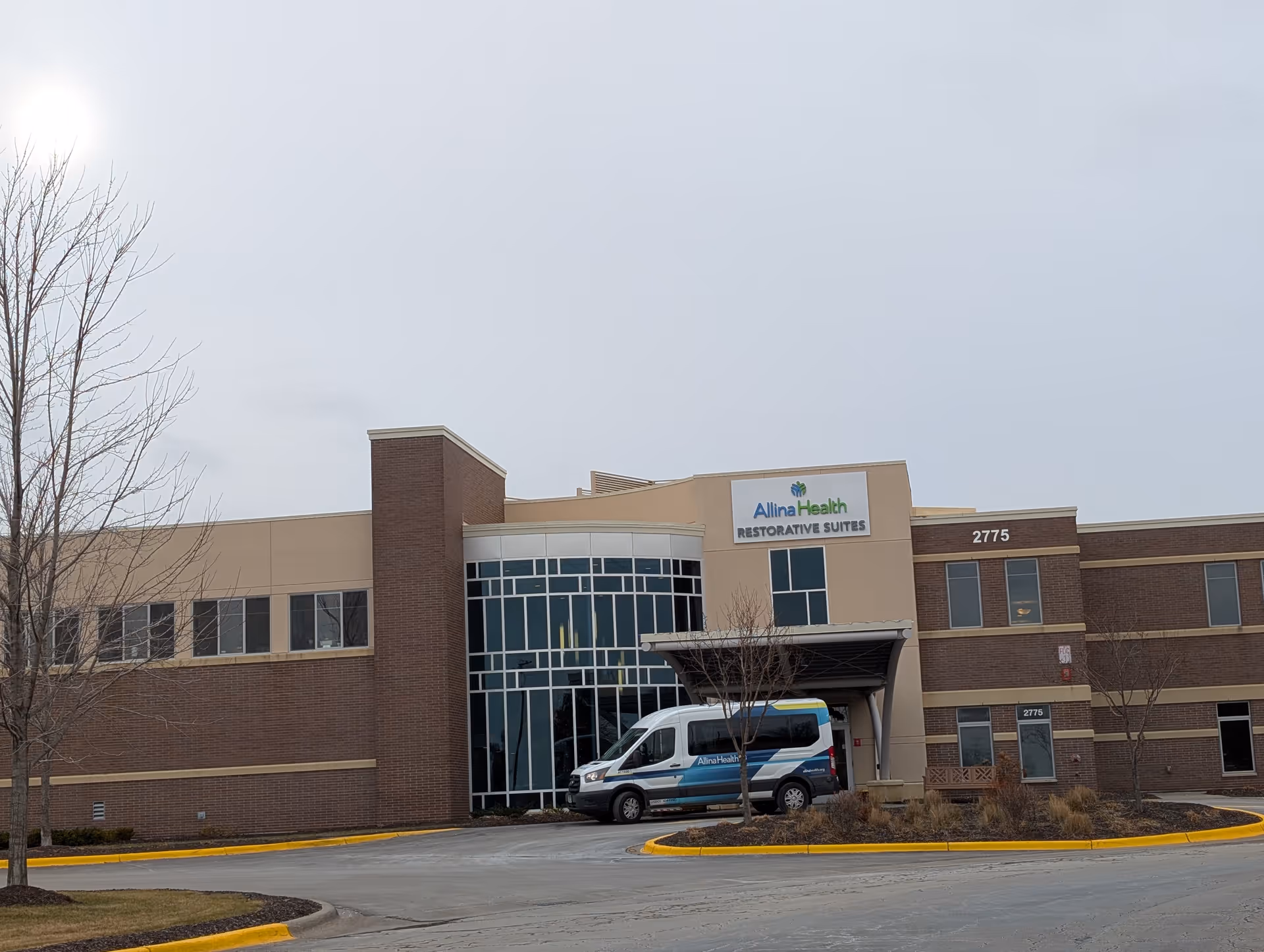 Exterior view of a two-story brick and beige building with large windows and a covered entrance. A white and blue Allina Health van is parked in front of the entrance. The building has a sign that reads 'Allina Health Restorative Suites' and the number 2775 is visible on the building.