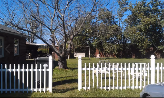 White picket fence surrounds a grassy outdoor area with trees, seating, and a swing set.