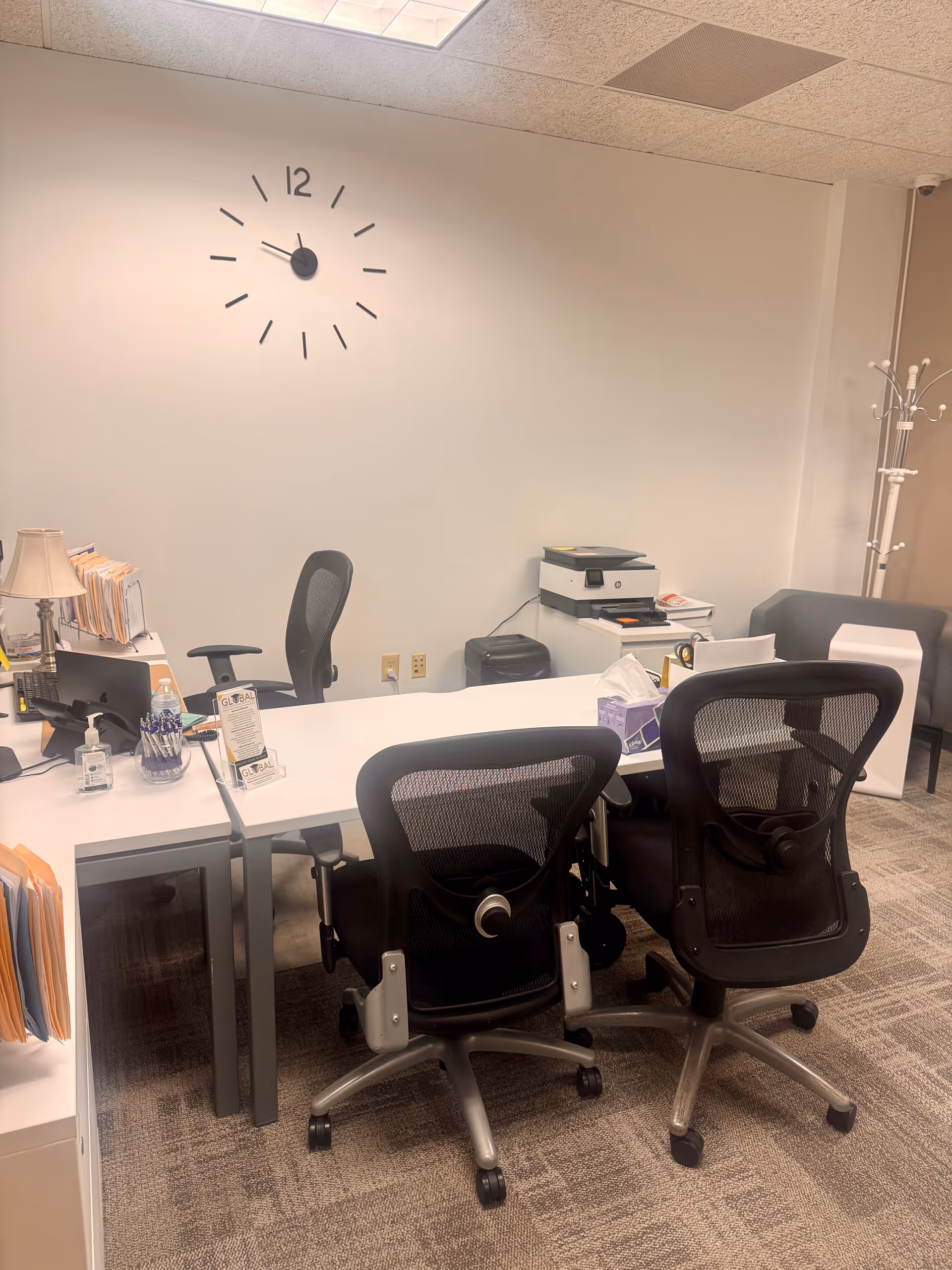Office workspace with white desk, two mesh chairs, printer, and wall clock.