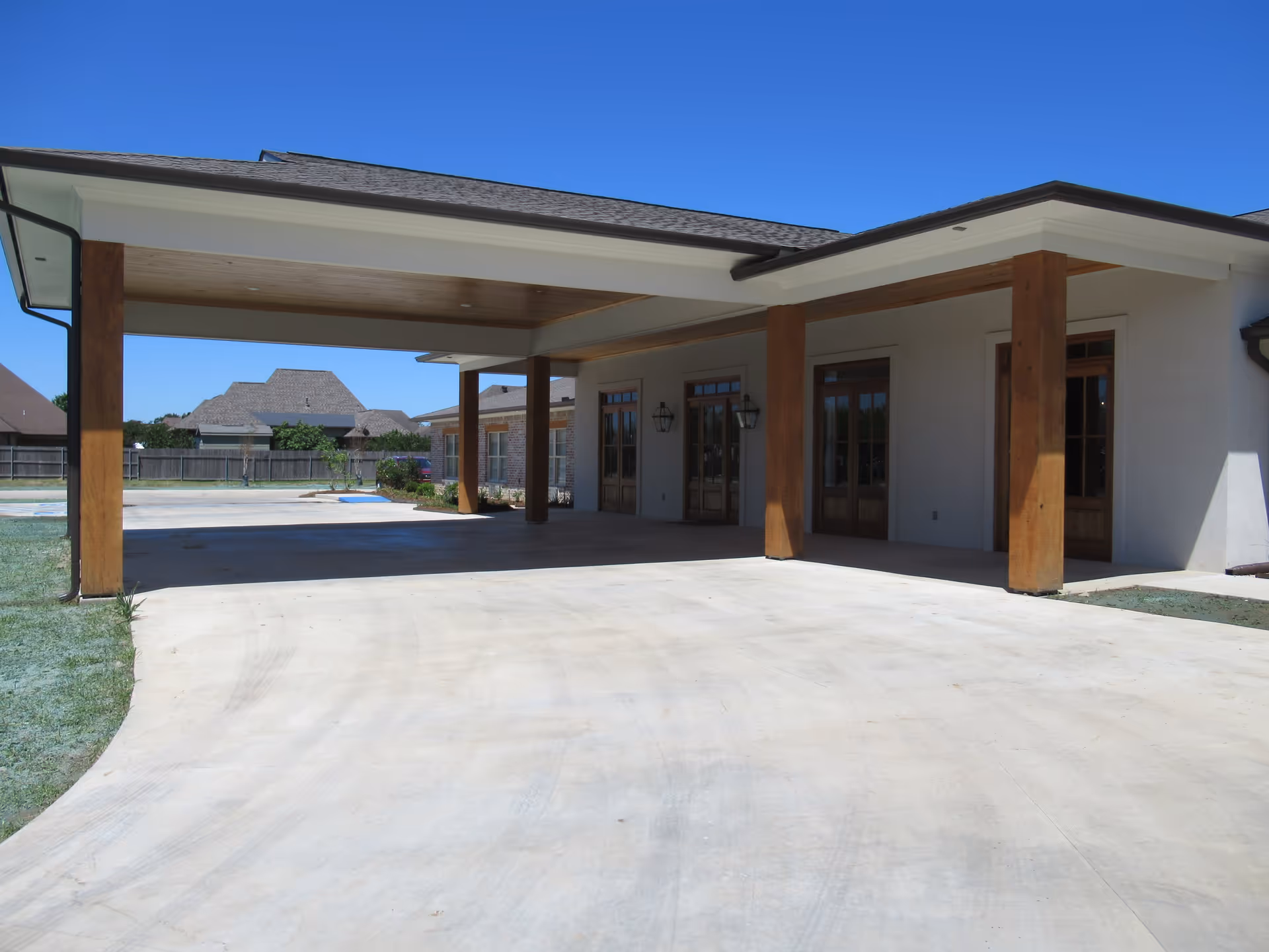 Covered driveway area with wooden pillars attached to a building with multiple wooden doors and wall-mounted lantern lights under a clear blue sky.
