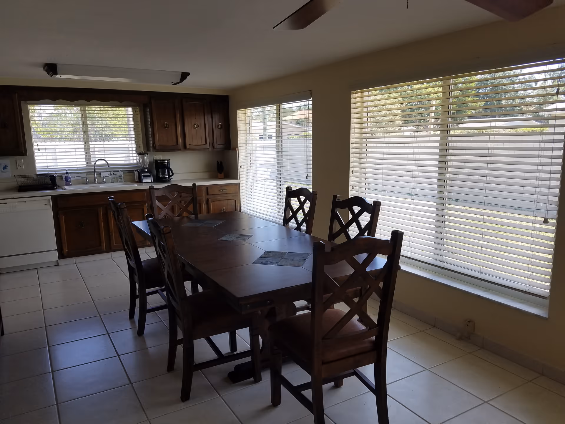 A kitchen and dining area with a wooden dining table and six matching chairs. The kitchen has wooden cabinets, a white dishwasher, a sink under a window with blinds, and appliances including a blender and coffee maker. Large windows with blinds let in natural light.