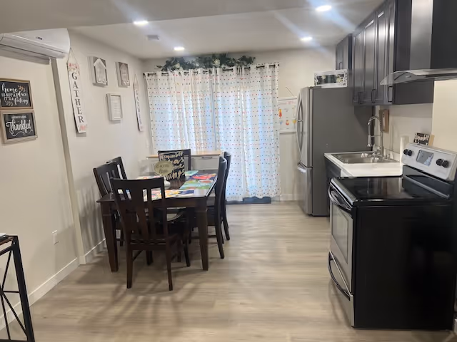 Bright interior kitchen and dining area with table, chairs, curtains, and stainless steel appliances.