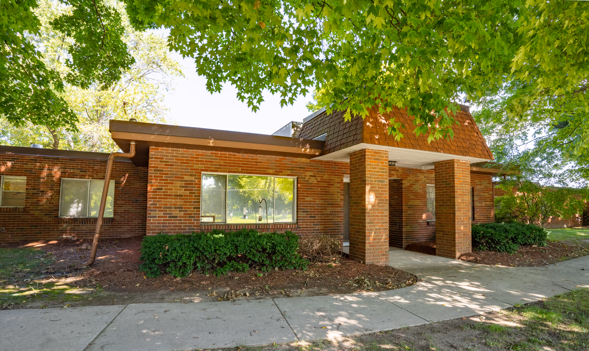 Exterior view of a single-story brick building with large windows and a covered entrance supported by brick columns. The building is surrounded by green trees and bushes, with a concrete sidewalk leading to the entrance.