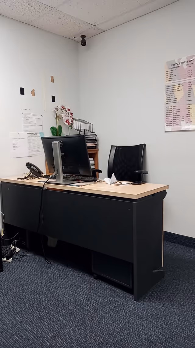 Office desk with monitor, phone, and paperwork in a small plain interior room.