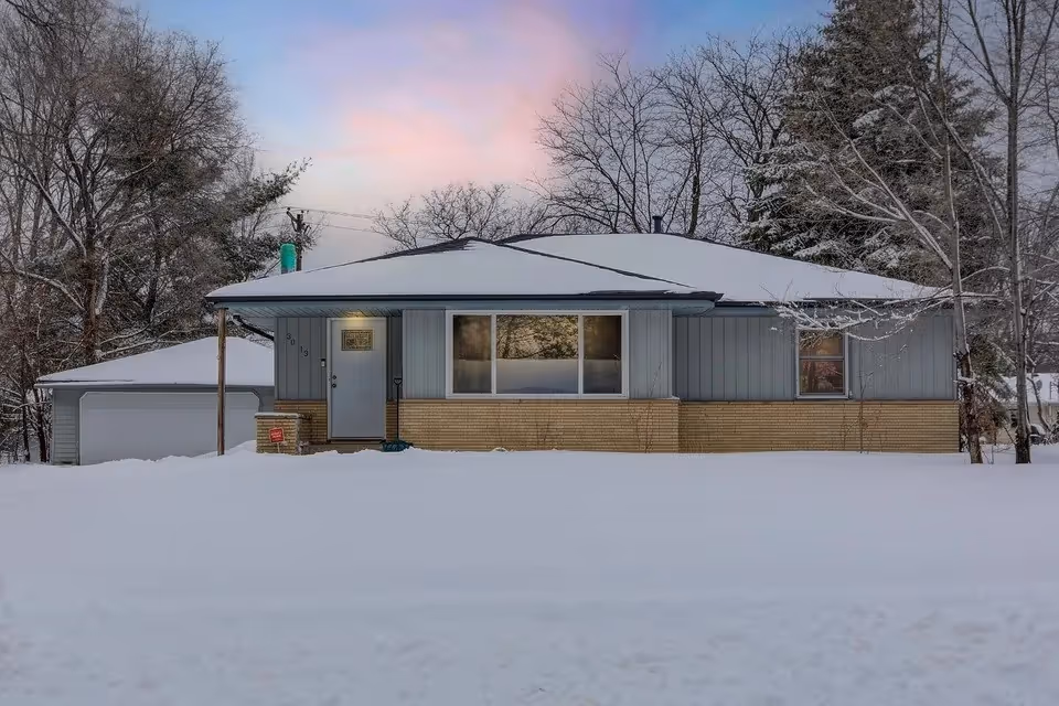 Snow-covered single-story building with an attached garage, bare trees, and pastel sunset sky.