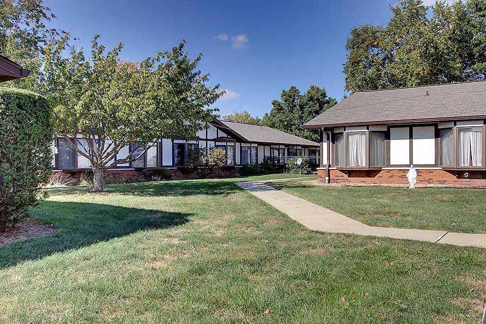 Courtyard with a grassy lawn, sidewalk, trees, and single-story brick buildings under a blue sky.