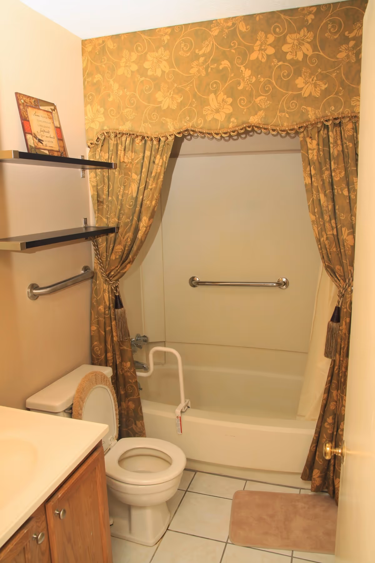 Bathroom with bathtub, toilet, sink, grab bars, and decorative patterned curtains in a senior facility.