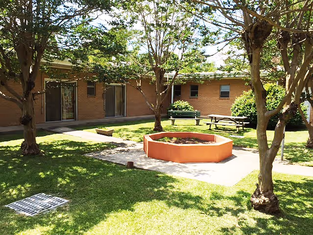 Green courtyard with trees, benches, a circular orange fire pit, and exterior building doors.