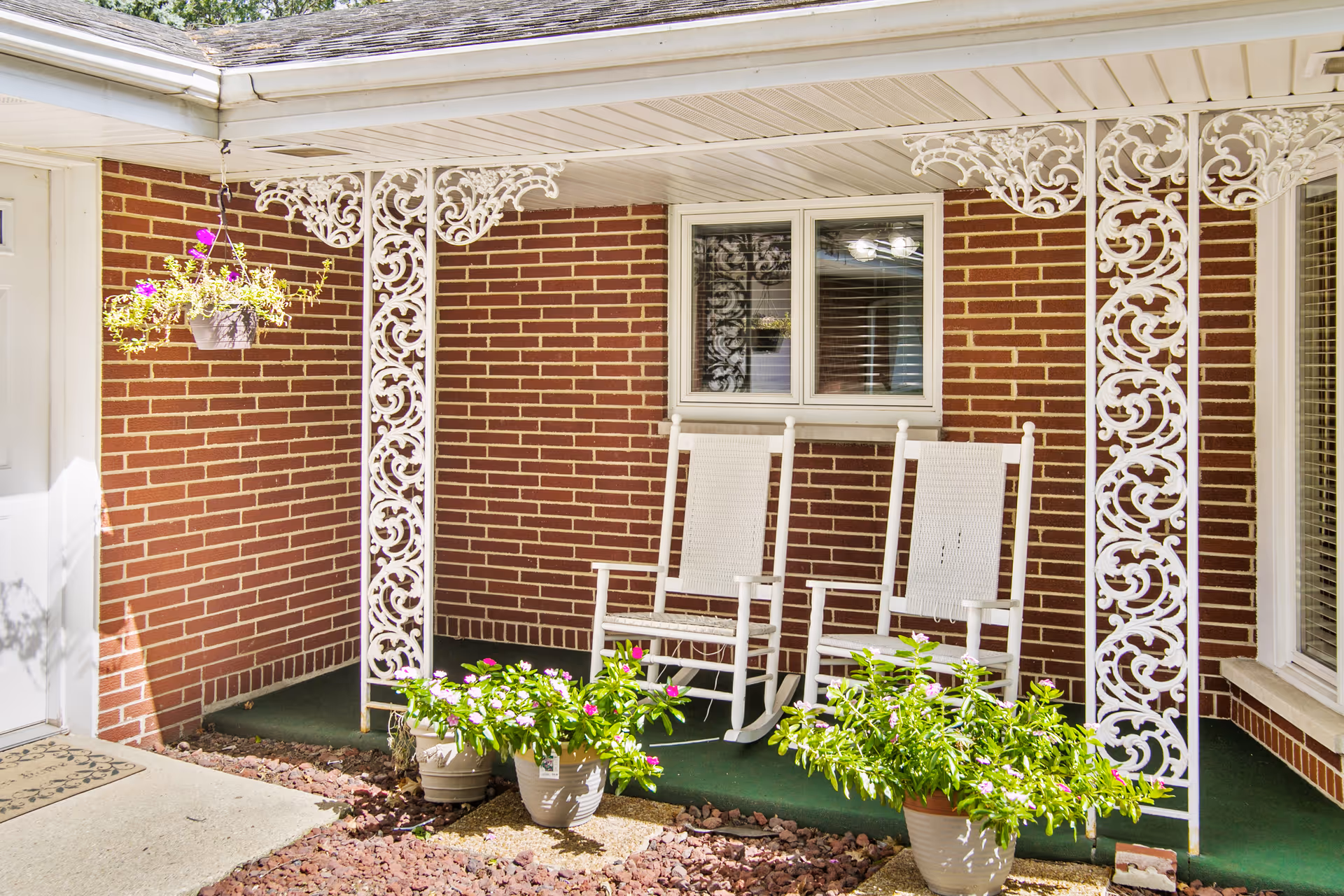 A small covered porch area with two white rocking chairs placed on a green carpet. The porch has decorative white wrought iron supports and a brick wall background. There are potted plants with green leaves and pink flowers on the ground, and a hanging flower pot with purple flowers on the left side.