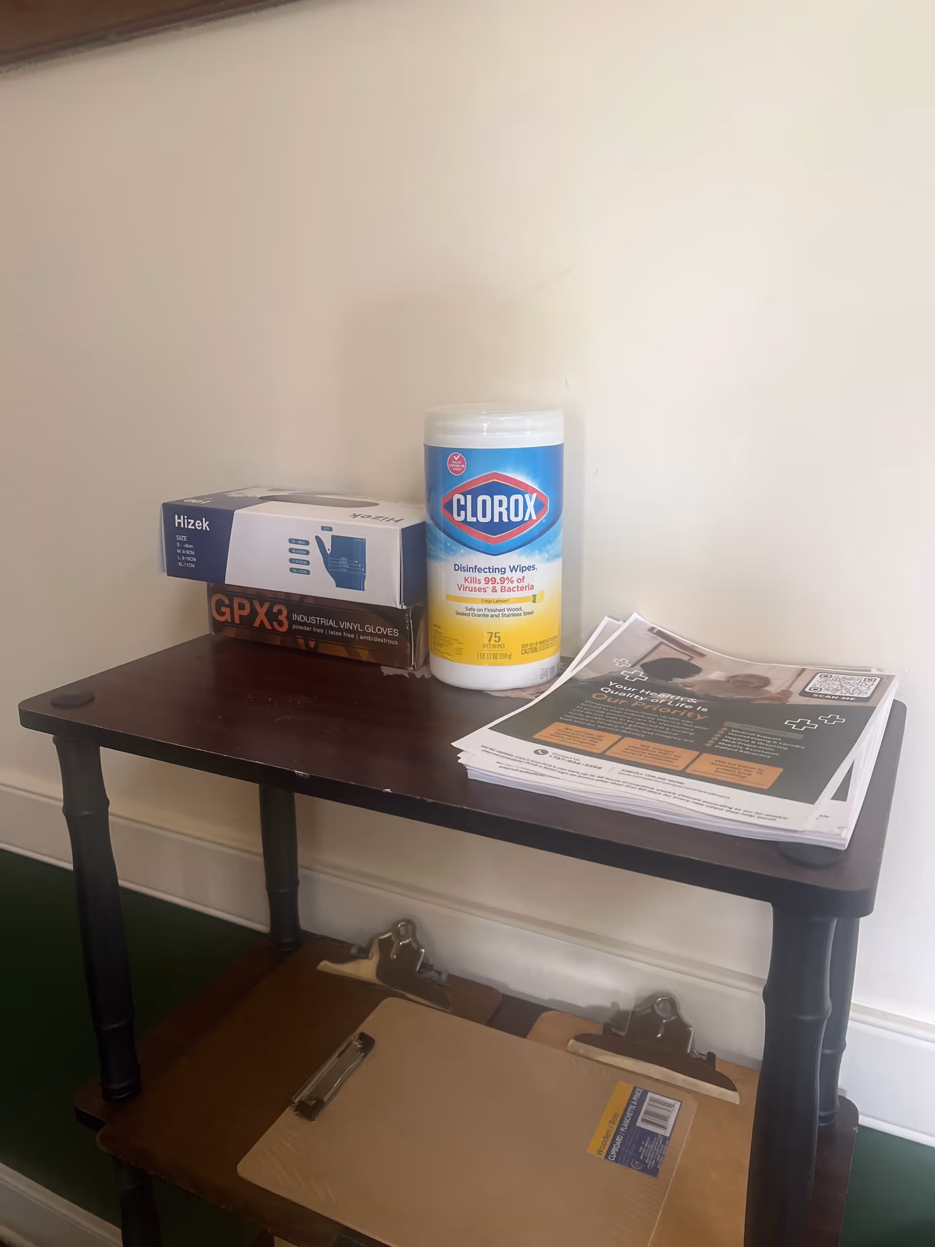 A wooden table against a white wall holds Clorox wipes, gloves, and a stack of papers.