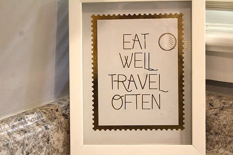 A framed decorative sign with a gold border and stylized text that reads 'EAT WELL TRAVEL OFTEN', placed on a granite countertop against a light-colored wall.