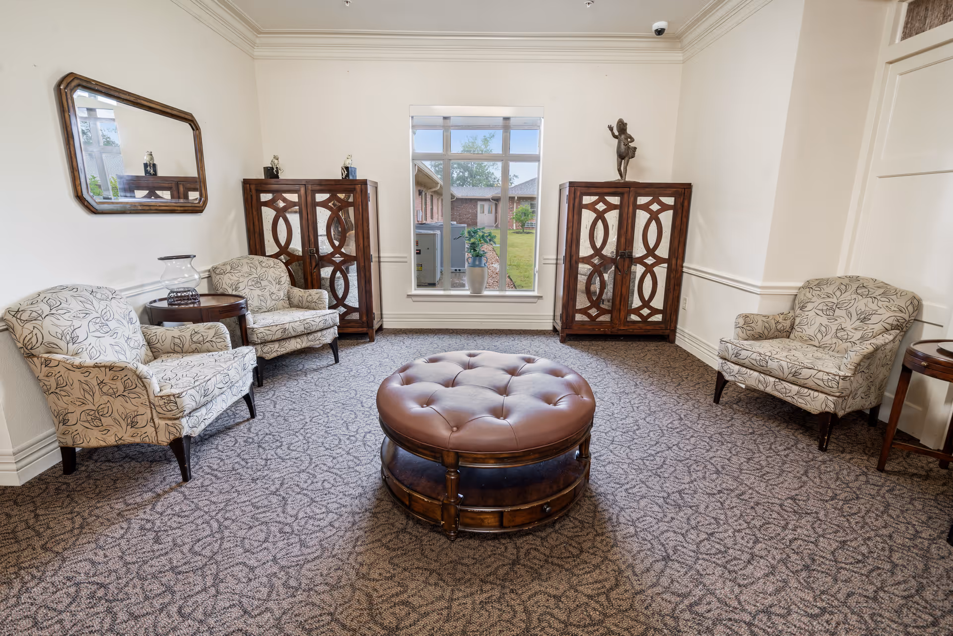 A cozy sitting area in a nursing and rehabilitation center featuring three patterned armchairs arranged around a large round tufted leather ottoman. The room has a patterned carpet, two wooden cabinets with decorative designs, a large window showing an outdoor view, a wall mirror, and small side tables with decorative items.