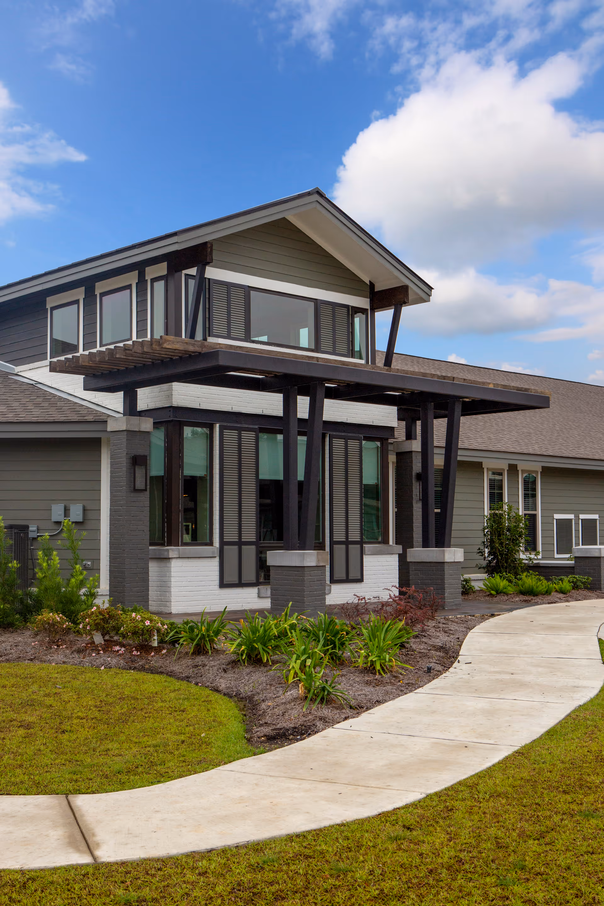 Exterior view of a modern senior living facility building with large windows, a covered entrance supported by dark beams, landscaped garden beds with green plants, and a curved concrete walkway leading to the entrance under a partly cloudy blue sky.