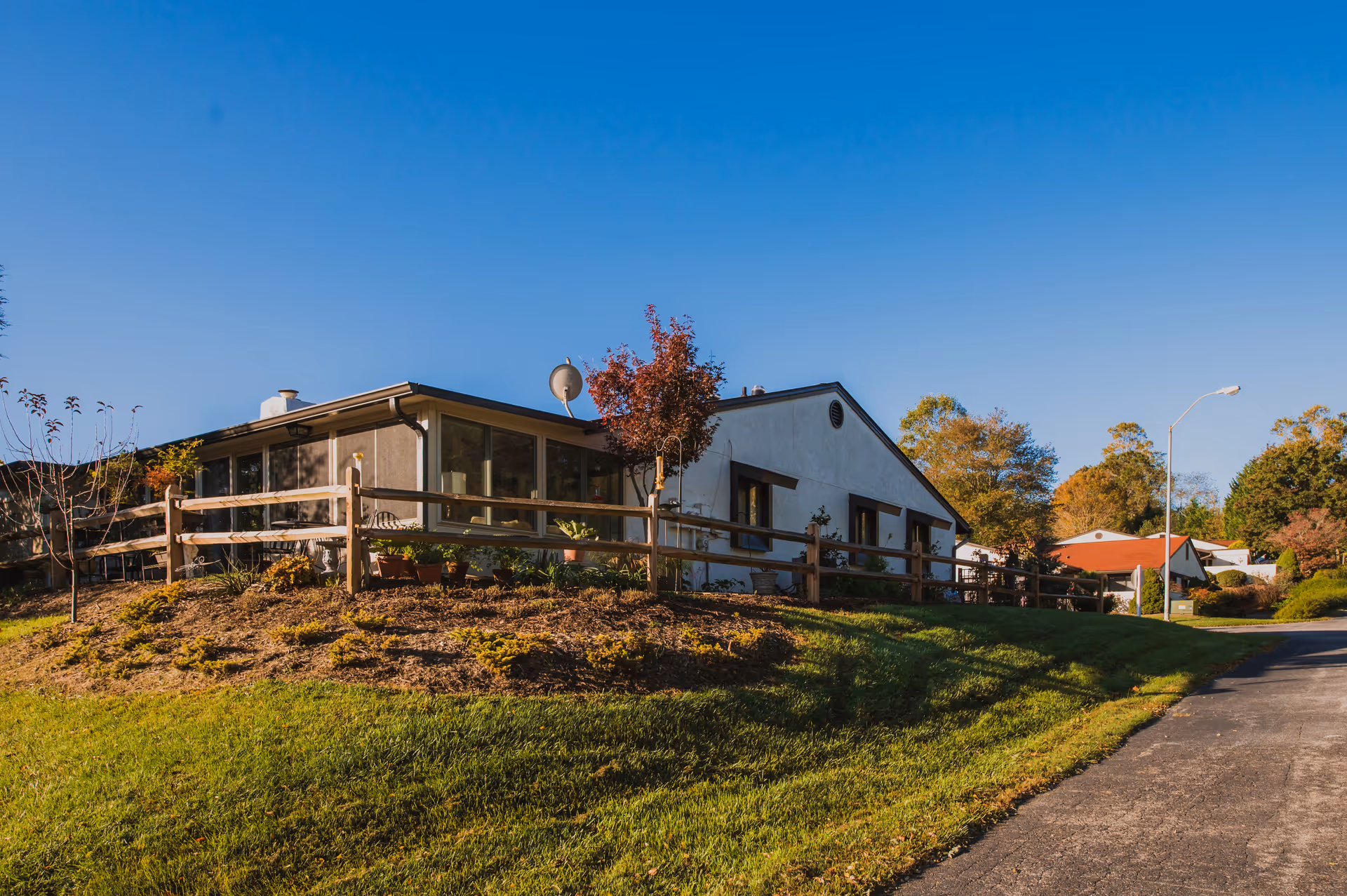 Exterior view of a single-story building with large windows, surrounded by a wooden fence and landscaping with small trees and plants under a clear blue sky.