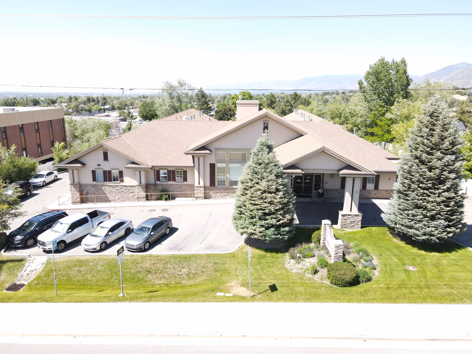 Exterior view of a single-story senior living facility building with a beige roof and stone accents. There are several parked cars in front of the building, two large evergreen trees flanking the entrance, and a well-maintained lawn with shrubs and a sign near the entrance. The background shows trees and distant mountains under a clear sky.
