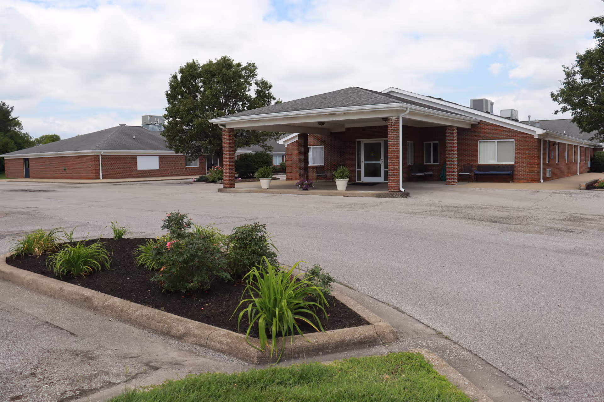 Exterior view of a single-story brick building with a covered entrance supported by brick columns, surrounded by a paved driveway and landscaped area with plants and shrubs under a partly cloudy sky.