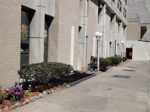 A sidewalk runs beside a beige brick building with trimmed hedges and flower beds.