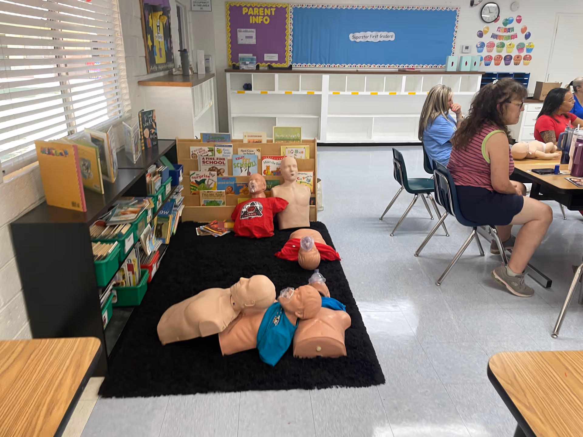 Indoor training room with CPR mannequins on a rug and adults seated nearby.