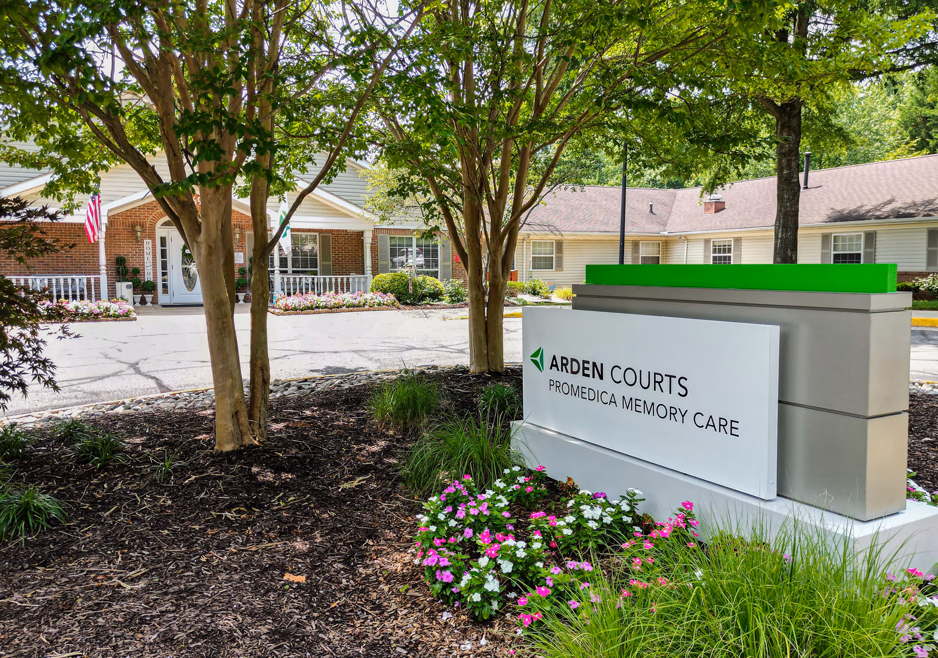 Entrance to Arden Courts ProMedica Memory Care with a prominent sign, trees, and landscaped flower beds in front of the building.