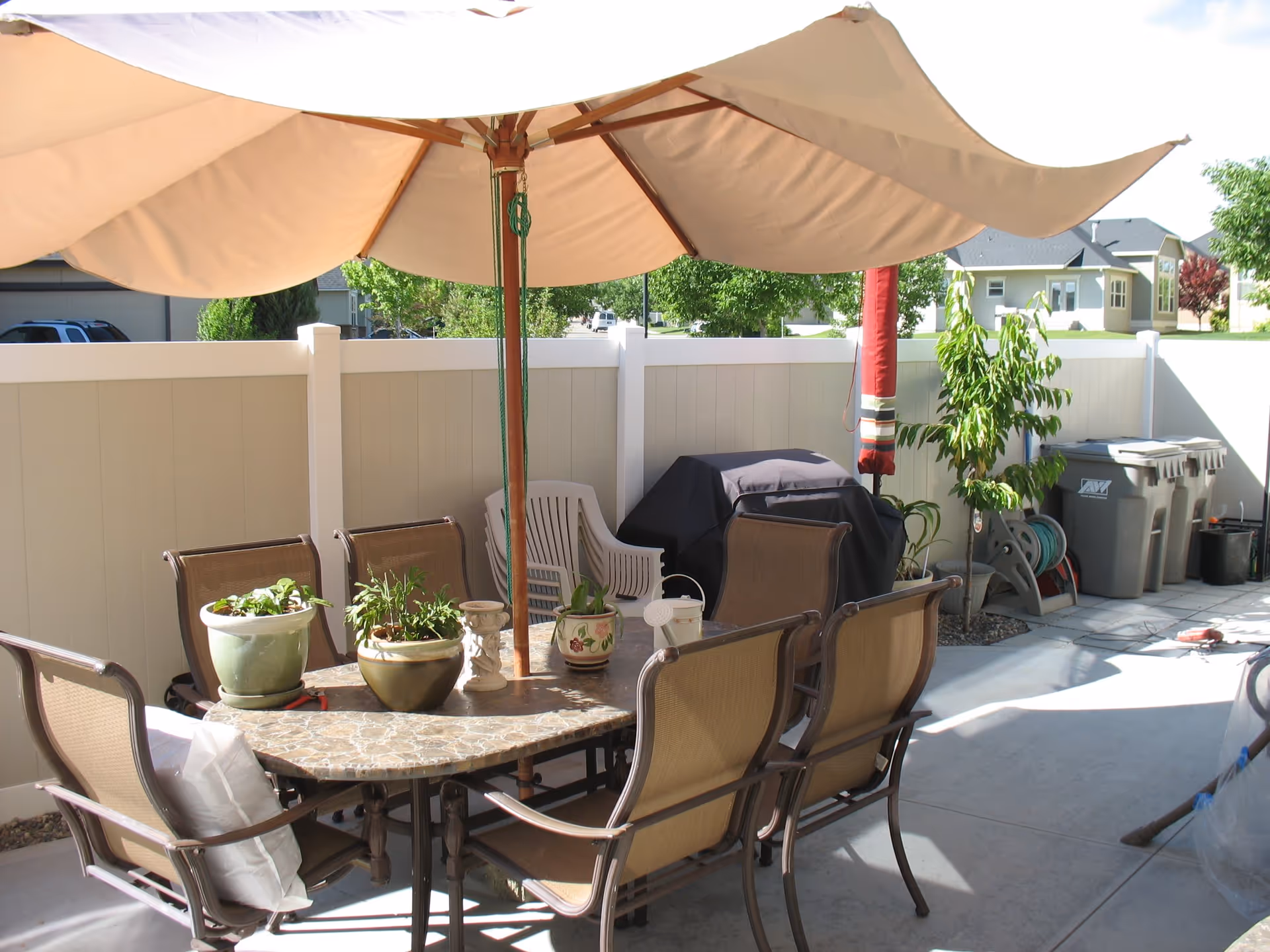 Outdoor patio area with a large beige umbrella shading a stone-top table surrounded by six brown mesh chairs. Several potted plants are placed on the table. In the background, there is a white privacy fence, a covered barbecue grill, stacked plastic chairs, a garden hose reel, and three trash bins. Residential houses and green trees are visible beyond the fence.
