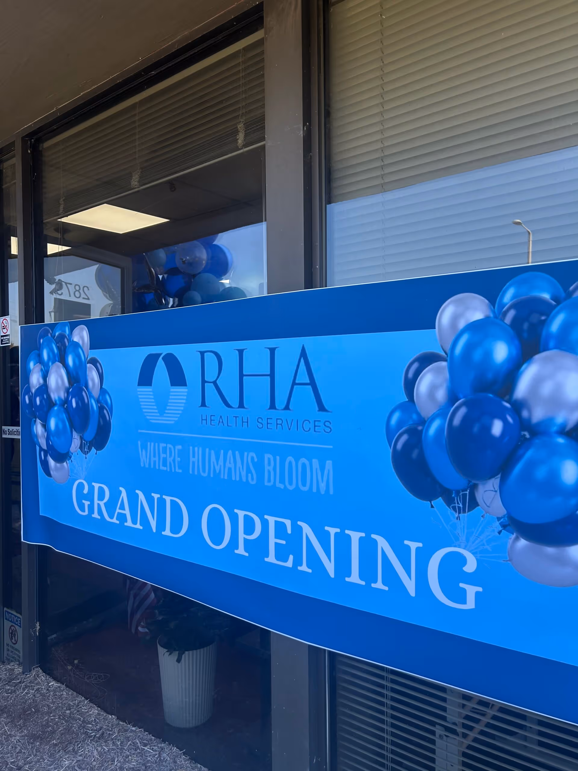 Blue banner reading 'GRAND OPENING' for RHA Health Services covers the storefront entrance with clusters of blue and silver balloons outside.