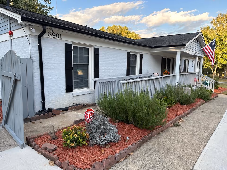 Single-story white brick senior care building with a wraparound porch, red mulch landscaping, and a flag.