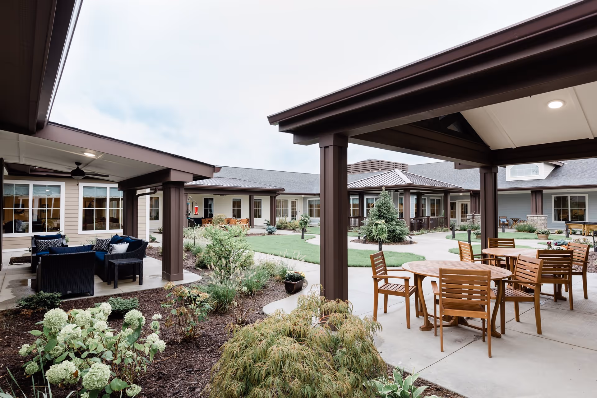 Outdoor courtyard area of Grand Brook Memory Care of Greenwood featuring covered seating areas with wooden tables and chairs, garden beds with various plants and shrubs, and a well-maintained lawn surrounded by the building's exterior walls and windows.